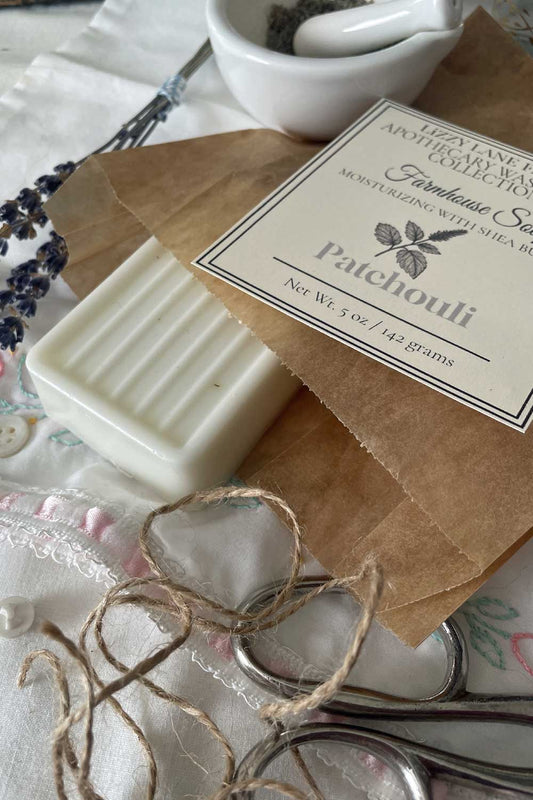 A creamy white Patchouli soap bar wrapped in kraft paper with twine and a classic label resting atop. Vintage lace and old buttons surround the scene, evoking a rustic, grounded farmhouse mood.