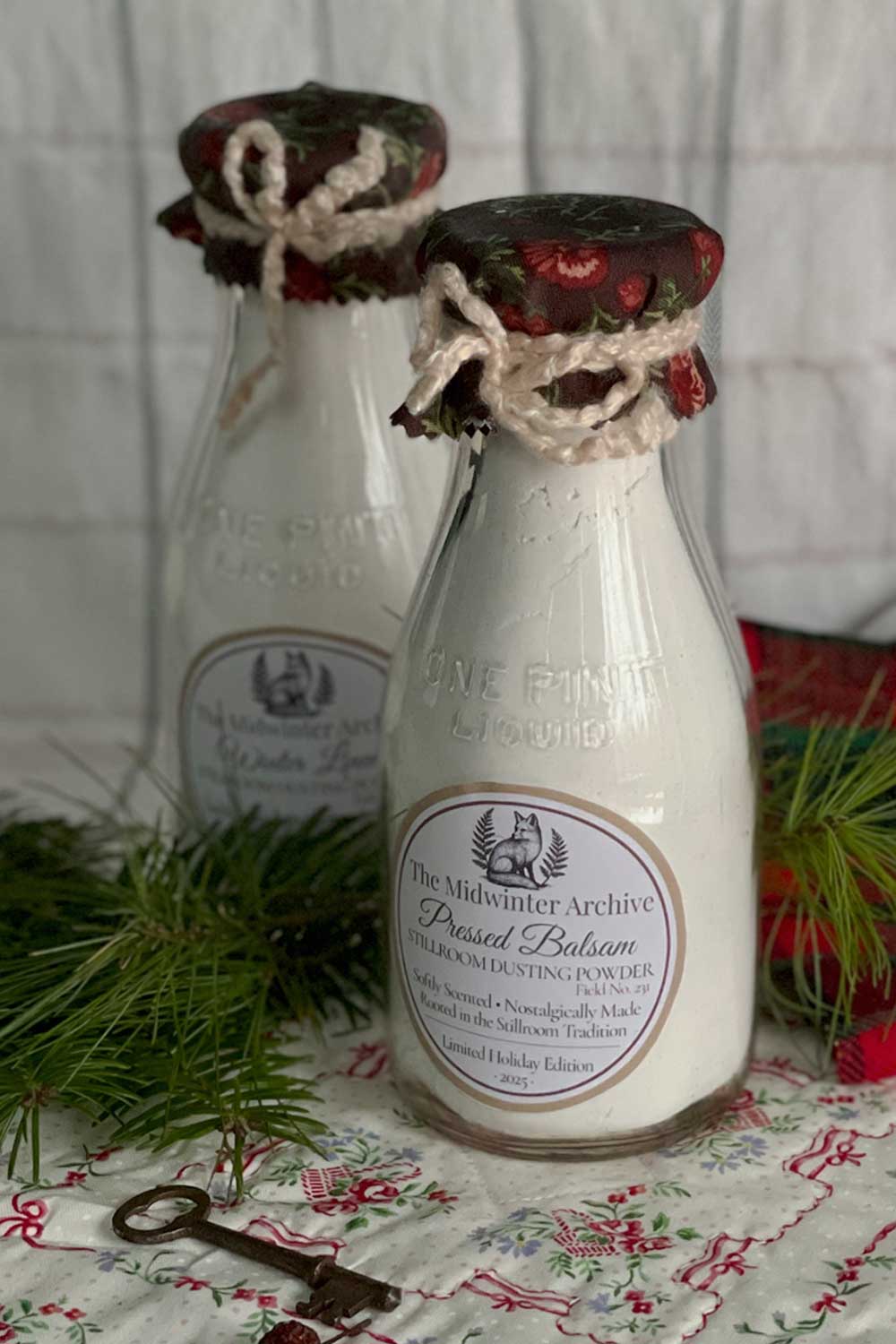 Pressed Balsam dusting powder in a collectible vintage milk bottle, cottagecore winter home décor with pine greenery and floral quilt background.
