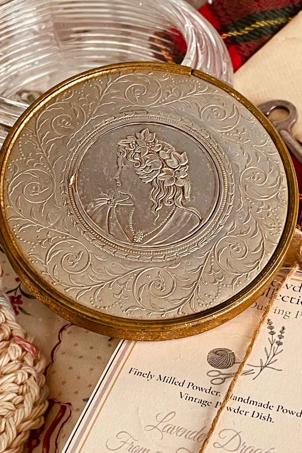 Close-up of a vintage powder dish lid featuring an embossed silver-and-gold cameo surrounded by ornate scrollwork, displayed with holiday greenery.