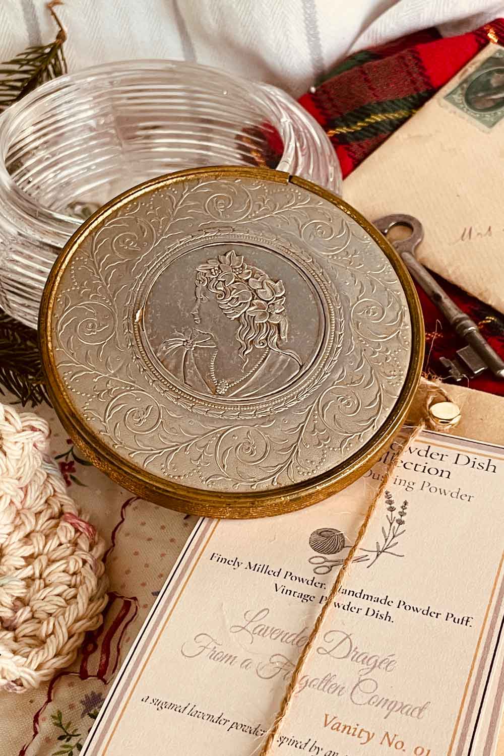 Close-up of a vintage powder dish lid featuring an embossed silver-and-gold cameo surrounded by ornate scrollwork, displayed with holiday greenery.
