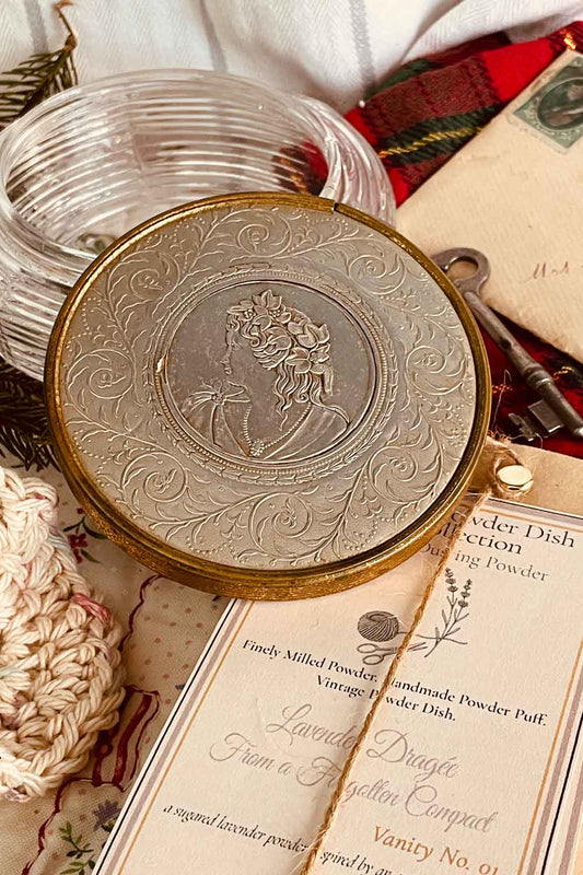 Close-up of a vintage powder dish lid featuring an embossed silver-and-gold cameo surrounded by ornate scrollwork, displayed with holiday greenery.