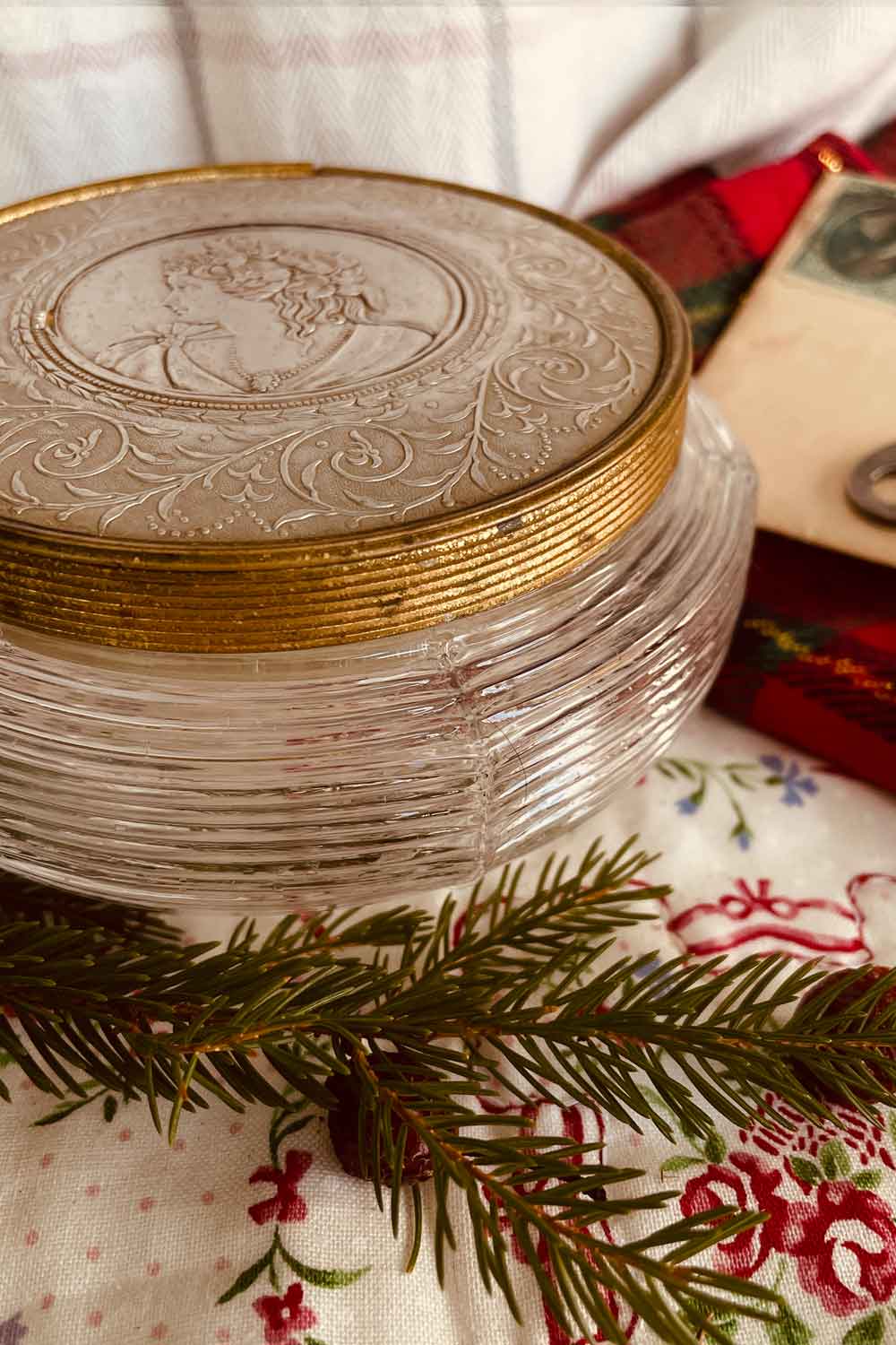 Vintage ribbed pressed-glass powder dish with embossed cameo lid and gold rim, styled on a floral cloth with evergreen sprig.