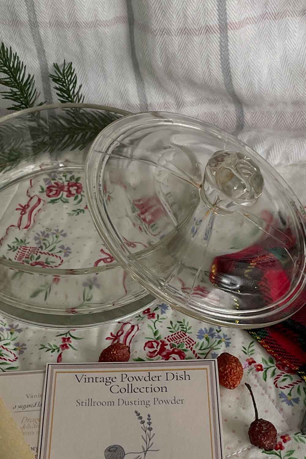 Vintage 1940s pressed-glass powder dish with rose-finial lid, displayed on a floral embroidered cloth with Stillroom Archive cards and Christmas greenery. Clear glass bowl and lid shown open to reveal condition and heirloom clarity.