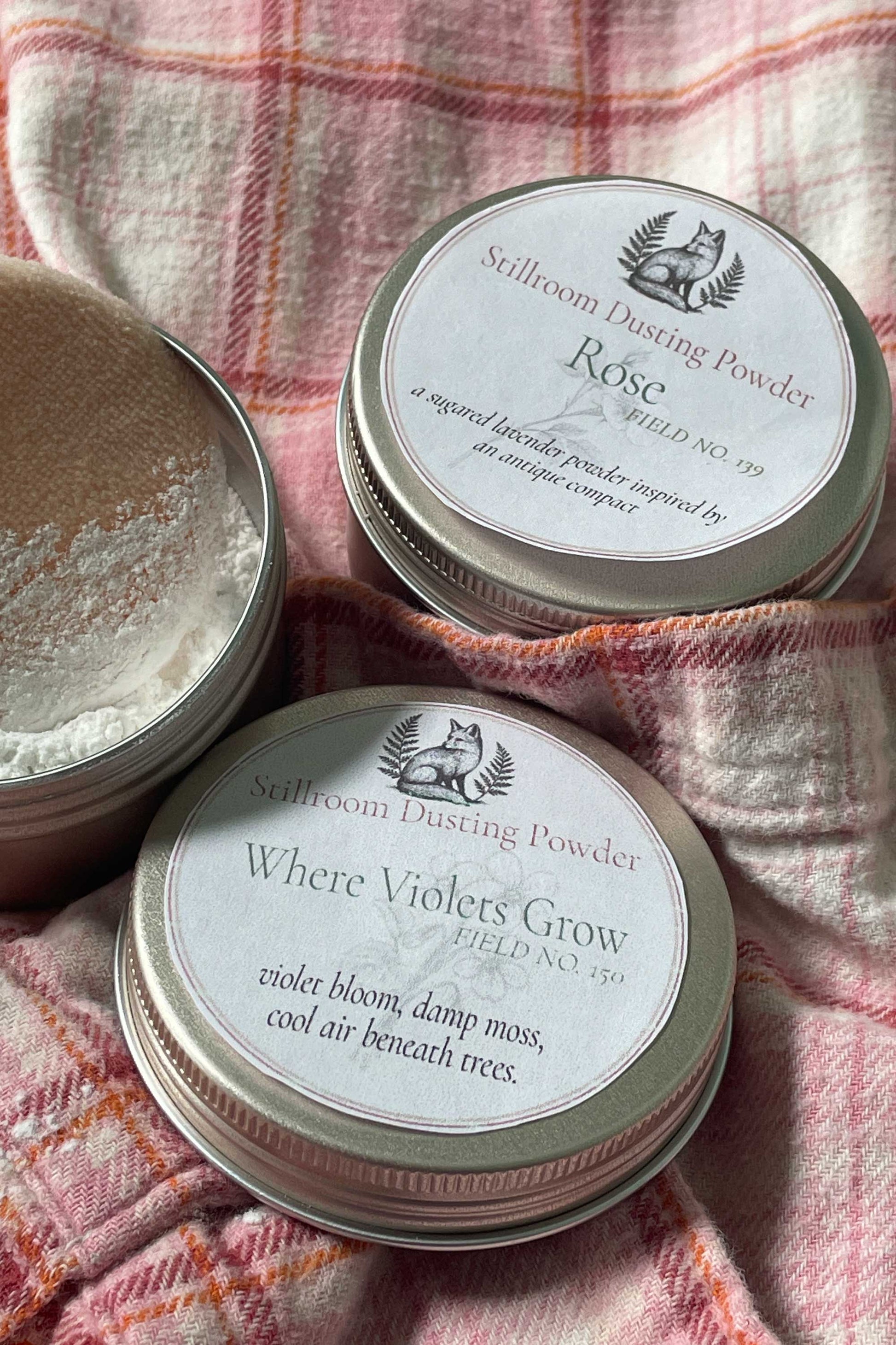 Three round metal tins of Stillroom Dusting Powder arranged on a pink plaid fabric background. One tin is open, showing white powder inside, while the other two show round white labels with black text.