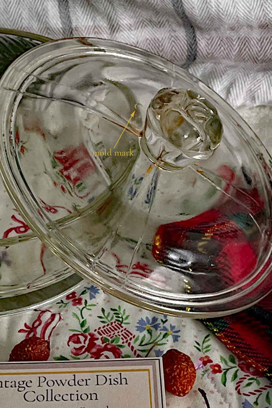 Close-up of vintage pressed-glass powder dish lid showing the sculpted rose finial and a visible original mold mark. Clear glass surface with radiating cut-glass lines, photographed on a floral quilt with winter Stillroom packaging.