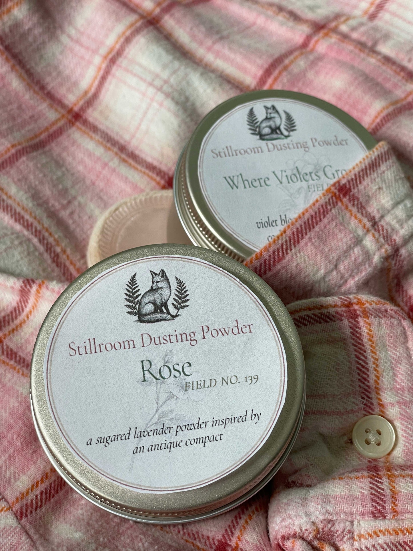 Two round metal tins of Stillroom Dusting Powder arranged on a pink plaid flannel shirt. The front tin is labeled 'Rose' and the back tin is labeled 'Where Violets Grow'.