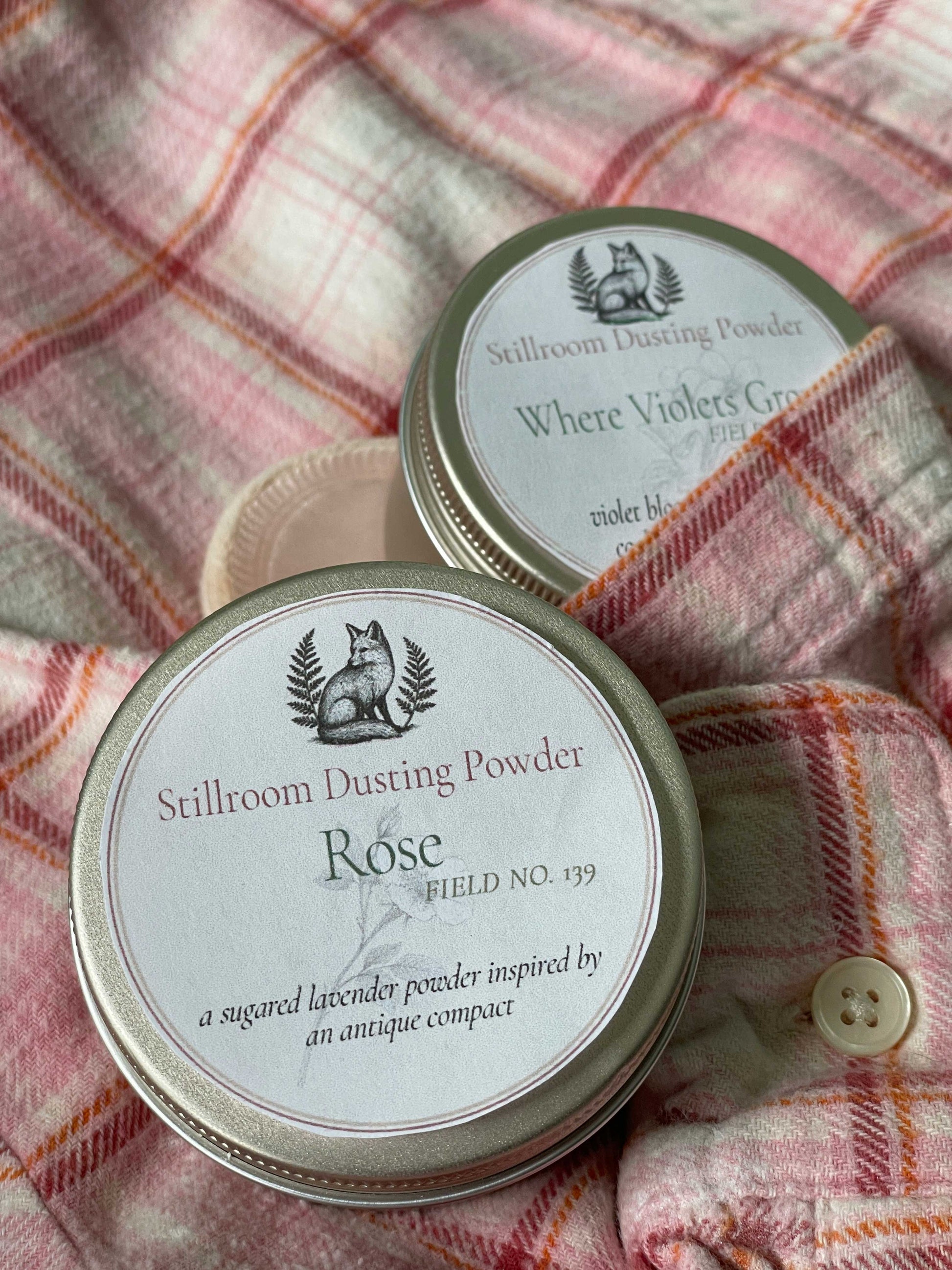 Two round metal tins of Stillroom Dusting Powder arranged on a pink plaid flannel shirt. The front tin is labeled 'Rose' and the back tin is labeled 'Where Violets Grow'.