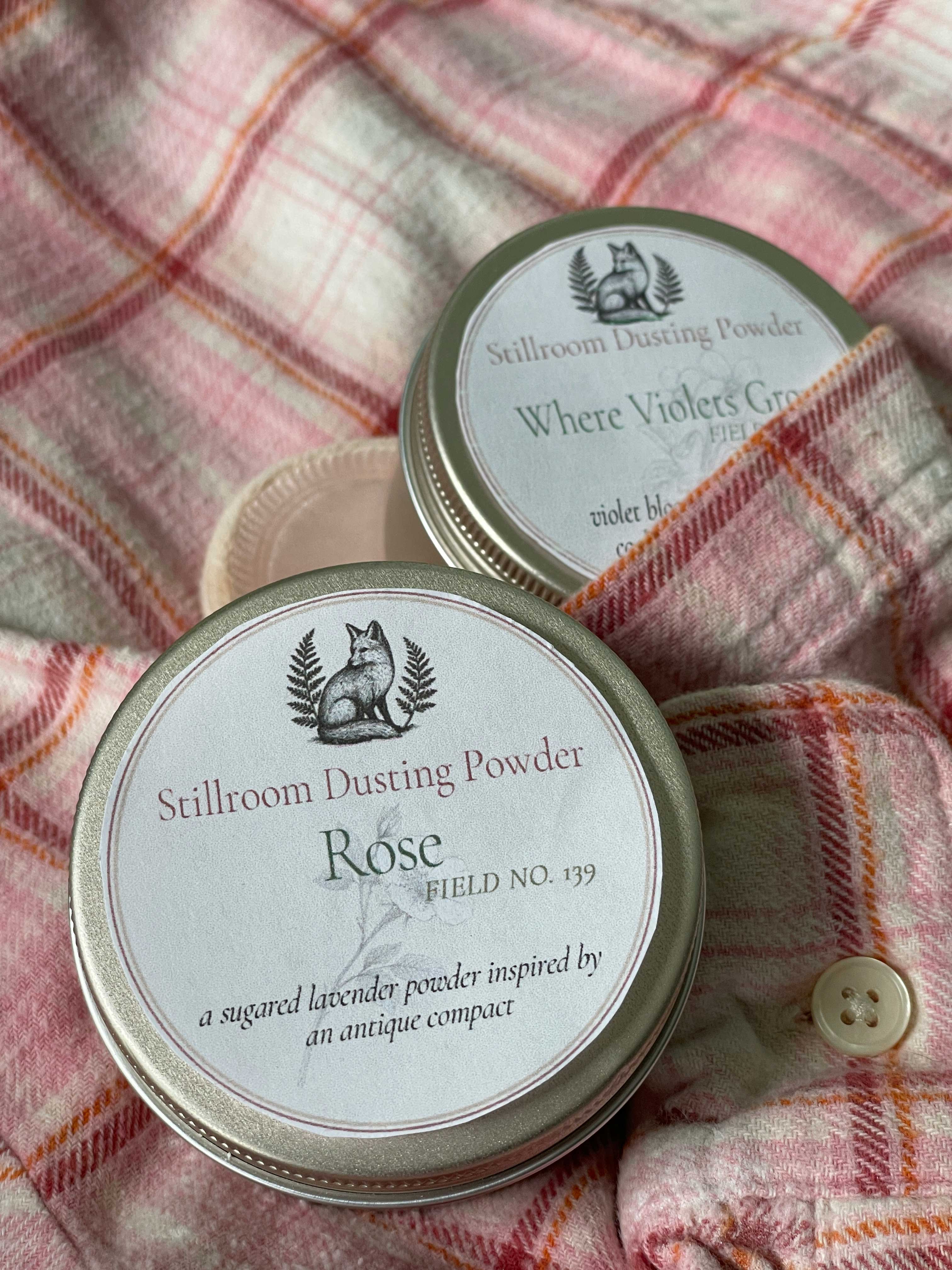 Two round metal tins of Stillroom Dusting Powder arranged on a pink plaid flannel shirt. The front tin is labeled 'Rose' and the back tin is labeled 'Where Violets Grow'.