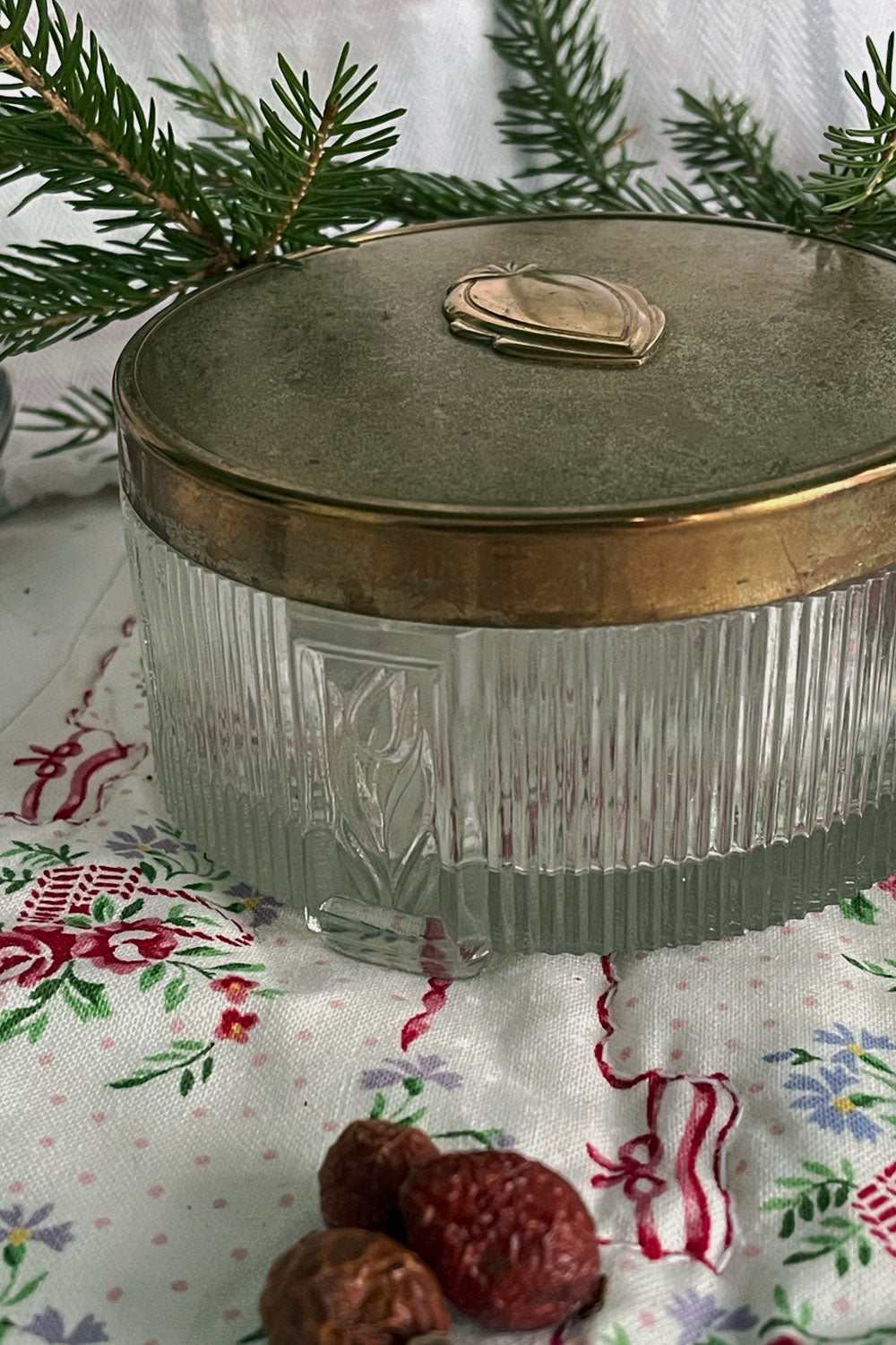 Vintage pressed-glass powder dish with raised floral design and brass Art Deco lid, photographed on a floral quilt with evergreen sprigs in soft natural light.