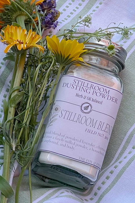 A glass powder shaker labeled 'Stillroom Dusting Powder' and 'Stillroom Blend' with a silver metal lid, styled with yellow and purple flowers on a green and white patterned fabric background.