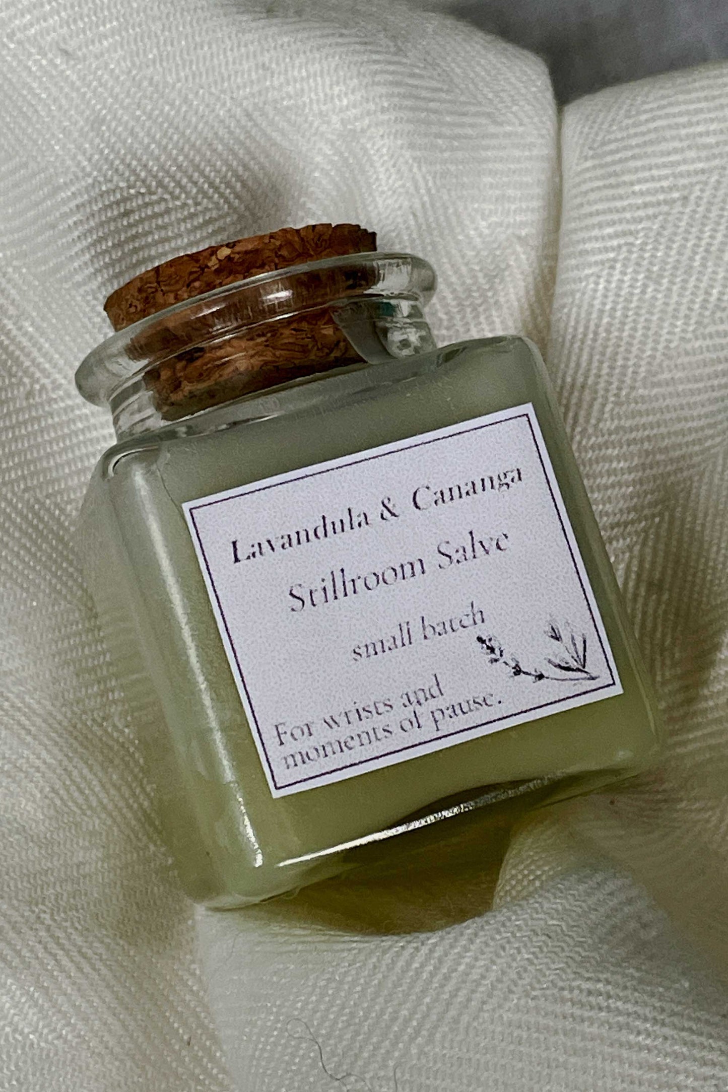 Small glass jar of 'Stillroom Salve' with a cork lid on a textured white fabric background