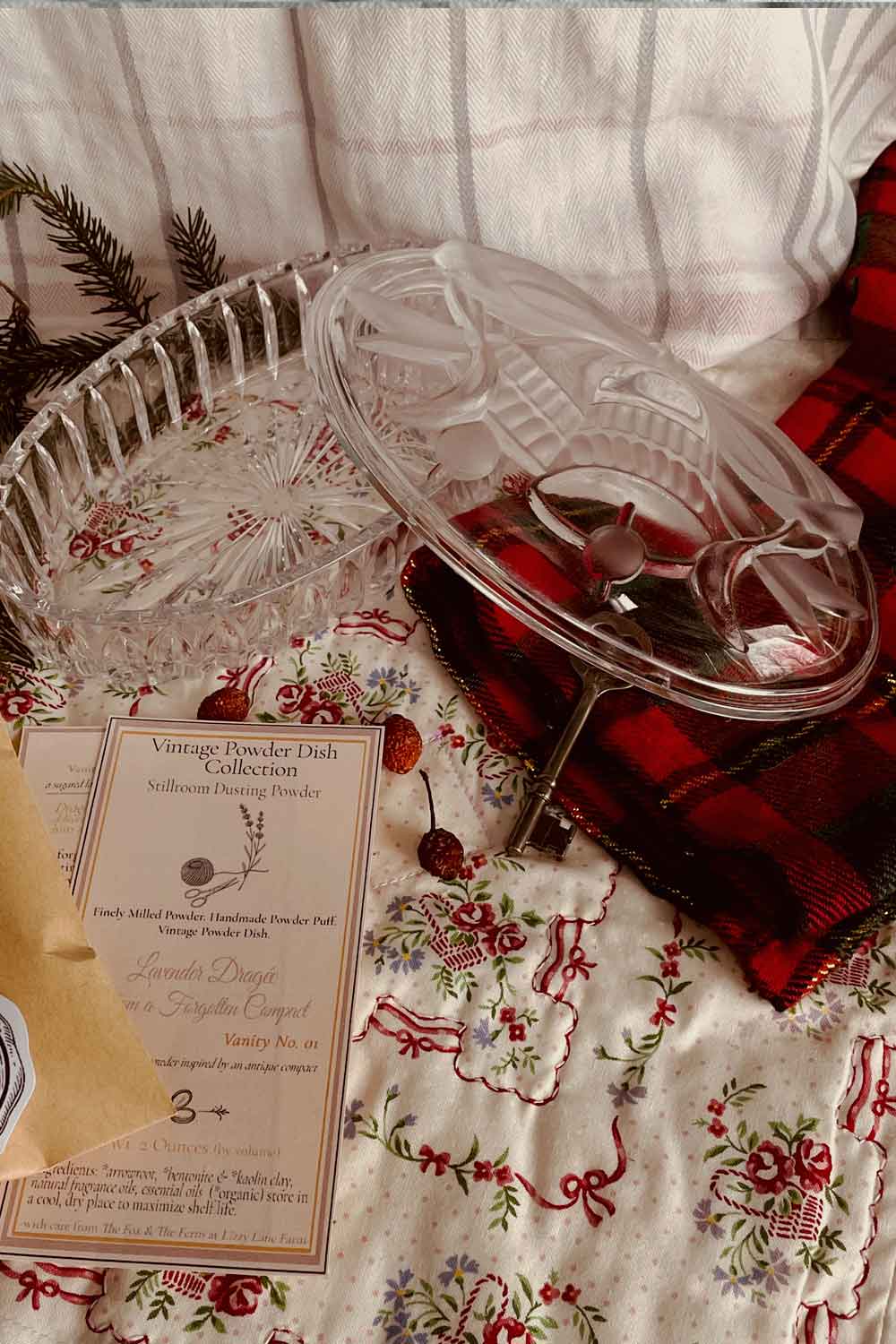 Vintage Studio Nova clear pressed-glass powder dish with Christmas bell motif, styled on a floral quilt with evergreen sprigs, Christmas plaid cloth, and Stillroom Archive cards for the Lavender Dragée powder.
