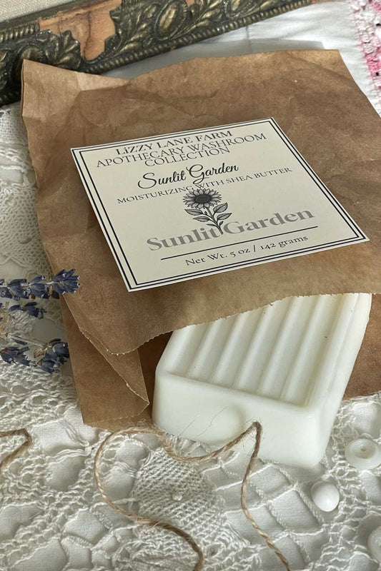 A creamy white bar of soap peeks from kraft paper wrapping, tied with twine and topped with a Sunlit Garden label. Vintage lace and scattered buttons frame the scene, with warm, golden sunlight softening the background.
