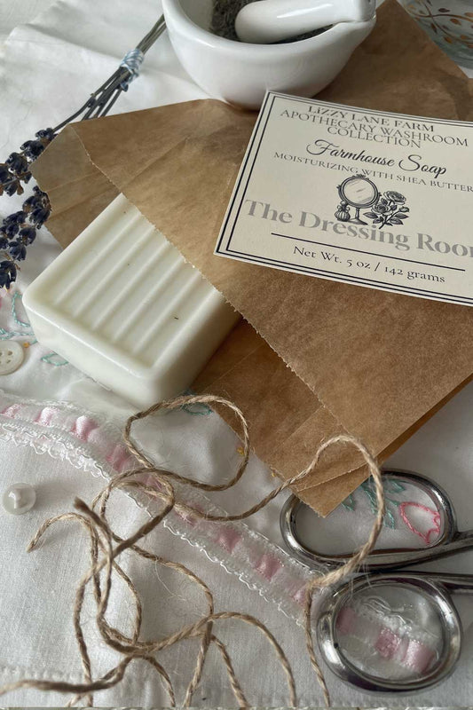 A creamy white bar of soap peeks out from kraft paper wrapping, tied with natural twine. The Dressing Room label rests atop, framed by delicate vintage lace and soft-hued buttons. Warm light spills across the scene, evoking a quiet, nostalgic moment.