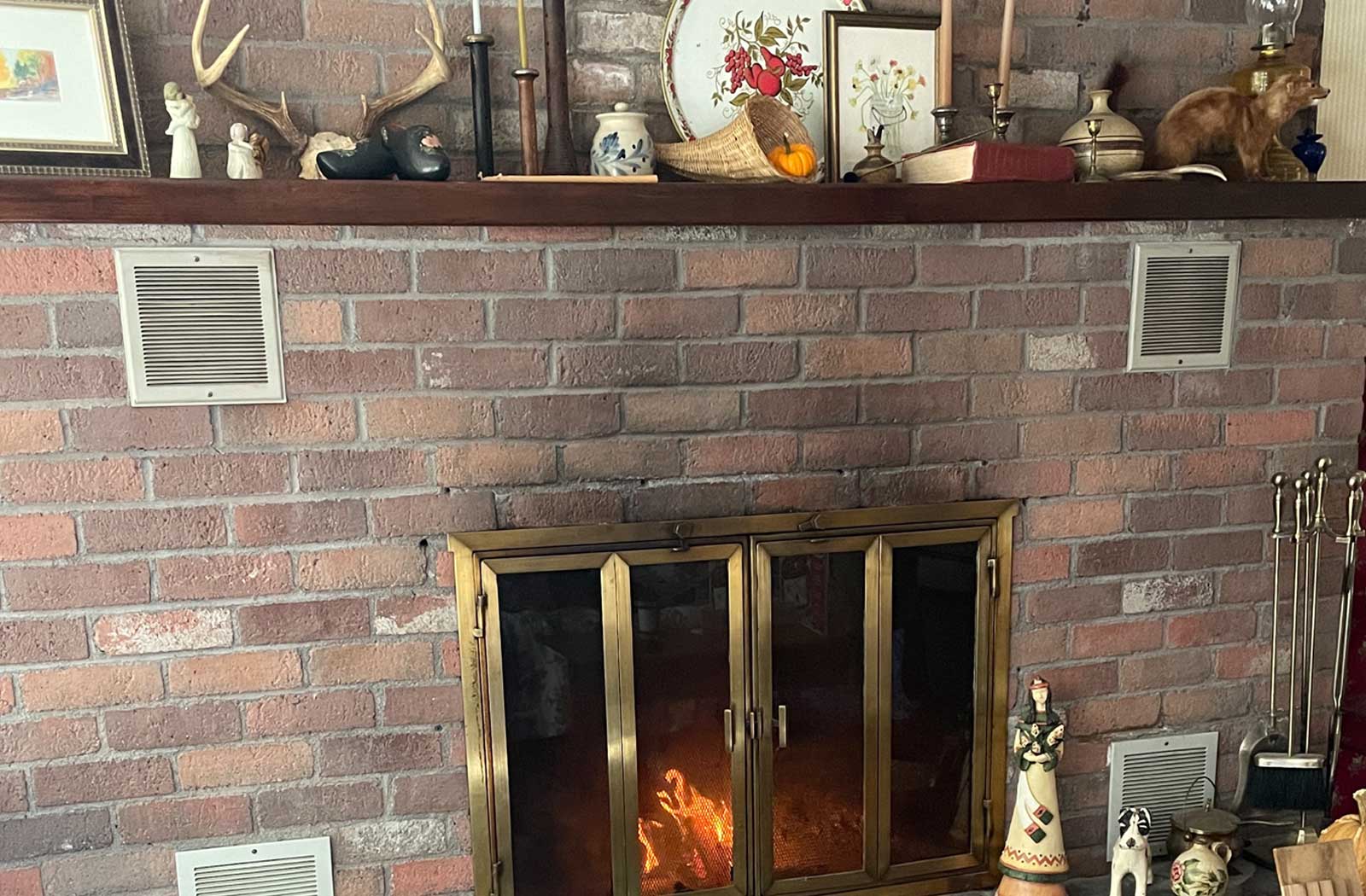 Brick farmhouse fireplace with brass doors, glowing fire, and a stillroom mantel decorated with candlesticks, crocks, books, and autumn accents — the Honeyed & Hearth collection banner, with the shop’s little weasel mascot tucked on the shelf.