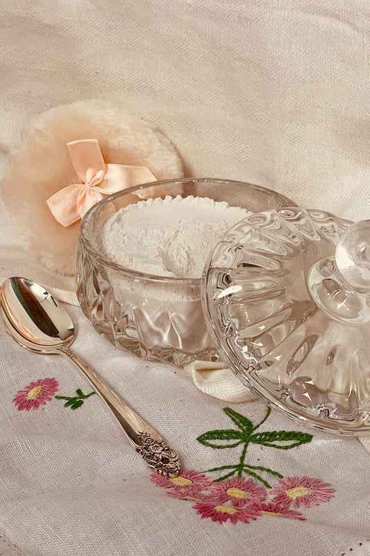 Stillroom-made powder dish set with glass vanity dish, lavender dusting powder, spoon, and puff arranged on linen