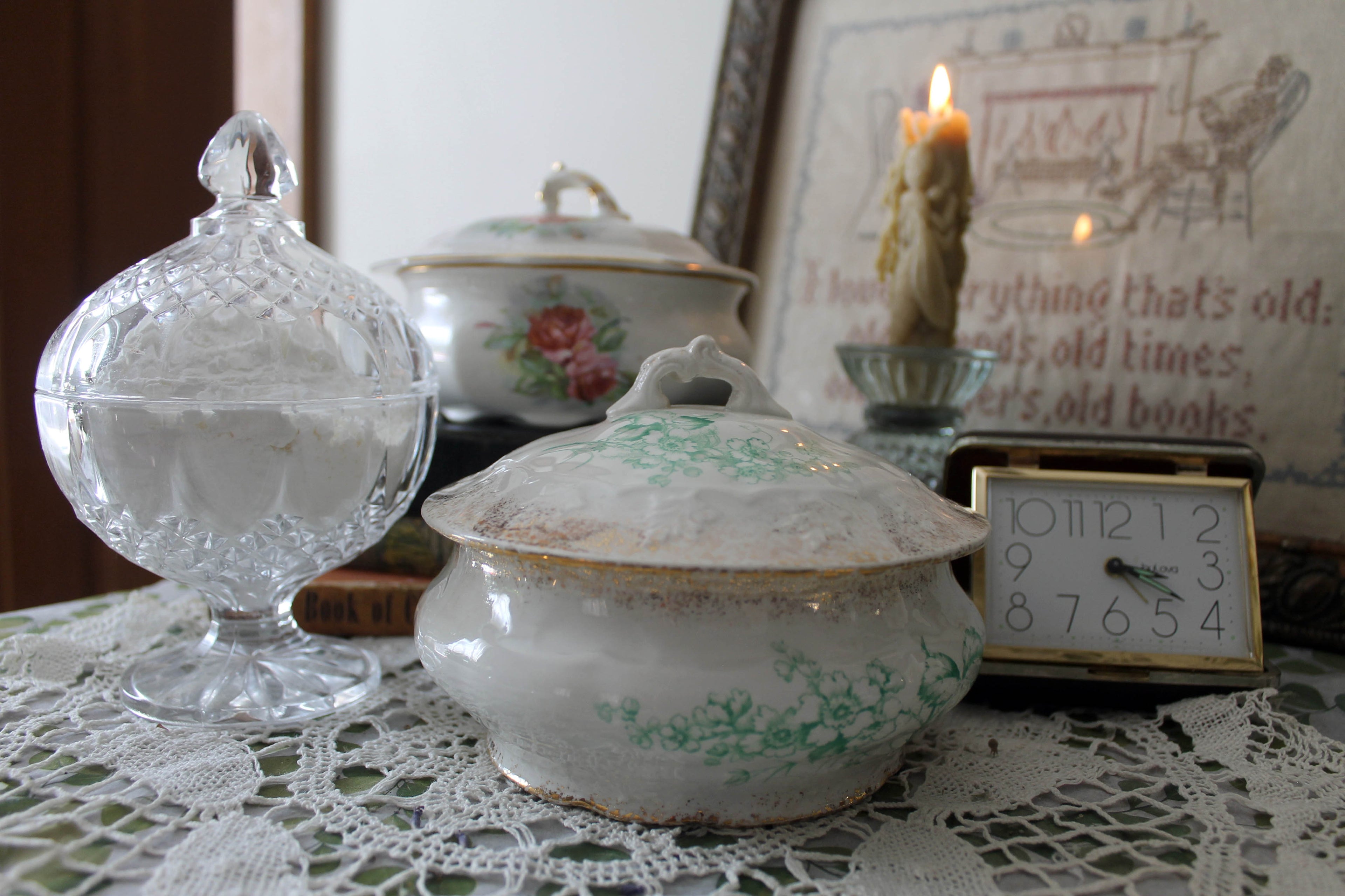 Vintage powder dishes, perfect to hold handcrafted dusting powder, resting on a romantic vintage dresser. Surrounded by delicate lace doilies, aged books, and soft, nostalgic charm, this collection evokes timeless elegance and a touch of vintage luxury.