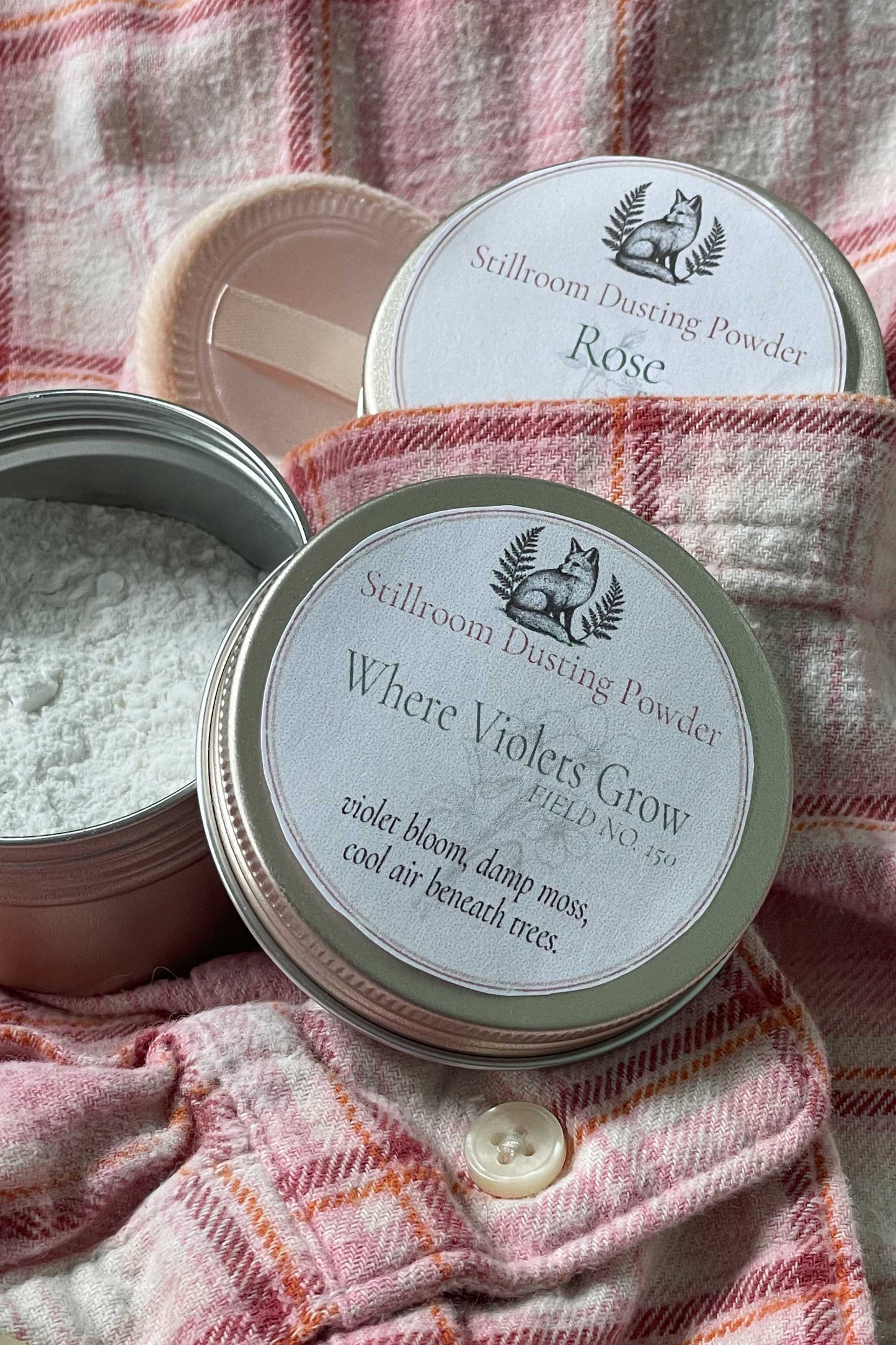 Two round metal tins of Stillroom Dusting Powder arranged on a pink plaid fabric background. One tin is open, showing white powder inside, and the other is closed with a printed lid label reading 'Where Violets Grow' and 'Róse'.
