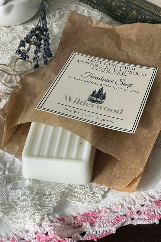 A creamy white soap bar wrapped in kraft paper with a twirl of natural twine. The Wilderwood label sits atop the package, surrounded by vintage lace and scattered buttons. Soft shadows and warm light evoke the quiet mystery of the woods beyond the farmhouse.