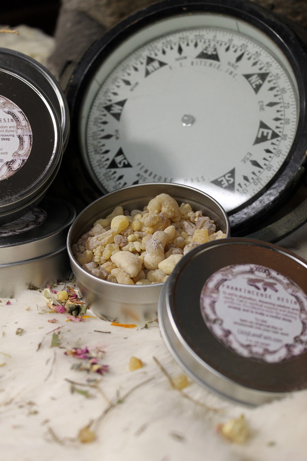 A silver metal tin filled with natural frankincense resin sits prominently in the foreground. The tin's sleek surface contrasts beautifully with the earthy, granular resin inside. In the background, a vintage compass adds a touch of rustic charm, enhancing the image with an old-world, adventurous vibe. The combination of the resin and compass evokes a sense of exploration and discovery, inviting you to embark on a sensory journey.