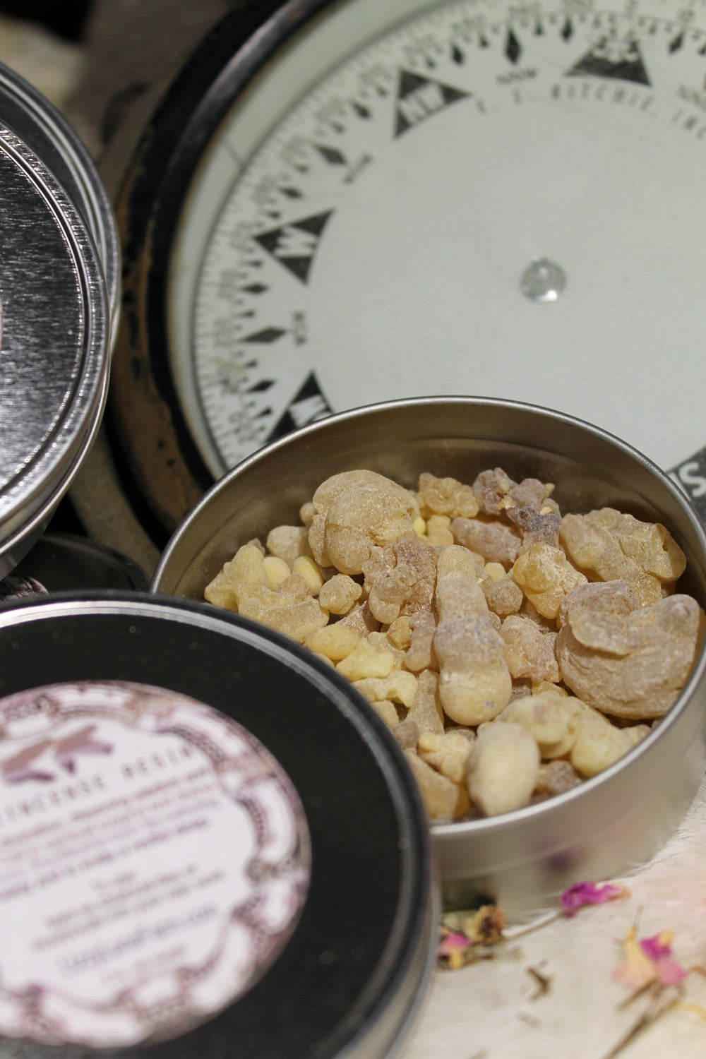 A silver metal tin filled with natural frankincense resin sits prominently in the foreground. The tin's sleek surface contrasts beautifully with the earthy, granular resin inside. In the background, a vintage compass adds a touch of rustic charm, enhancing the image with an old-world, adventurous vibe. The combination of the resin and compass evokes a sense of exploration and discovery, inviting you to embark on a sensory journey.