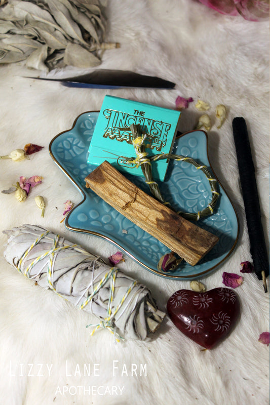 A vibrant teal Hamsa hand smudge plate takes center stage in this beautifully arranged smudge kit. Intricate details and a gold-tone rim enhance the spiritual design, while surrounding it are the thoughtfully curated supplies: a mini white sage smudge stick, a braided sweet grass bundle, a palo santo stick, hand-rolled Styrax incense, a hand-carved soapstone heart, a Chinese Qing Dynasty coin charm, and rain-scented incense matches. All these items are neatly packaged in a rustic burlap pouch, ready to brin